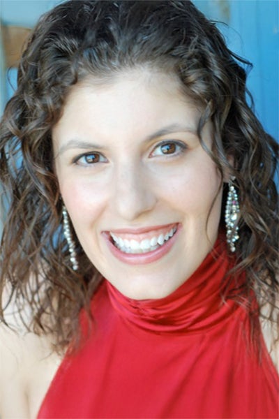 Close-up of a smiling woman with curly brown hair, wearing a red sleeveless top and dangling earrings.