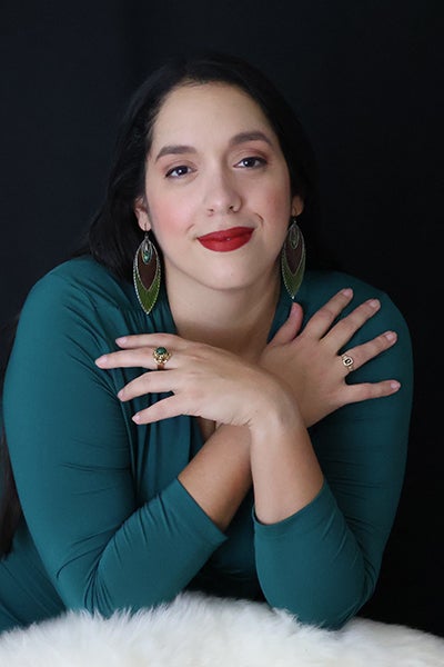 Portrait of a woman with dark hair in a teal top, smiling, arms crossed over her chest, against a dark background.