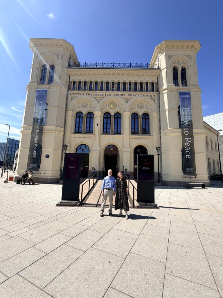 Two adults stand in front of the Nobels Fredsgssenter/Nobel Peace Center building with arched windows and banners on either side.