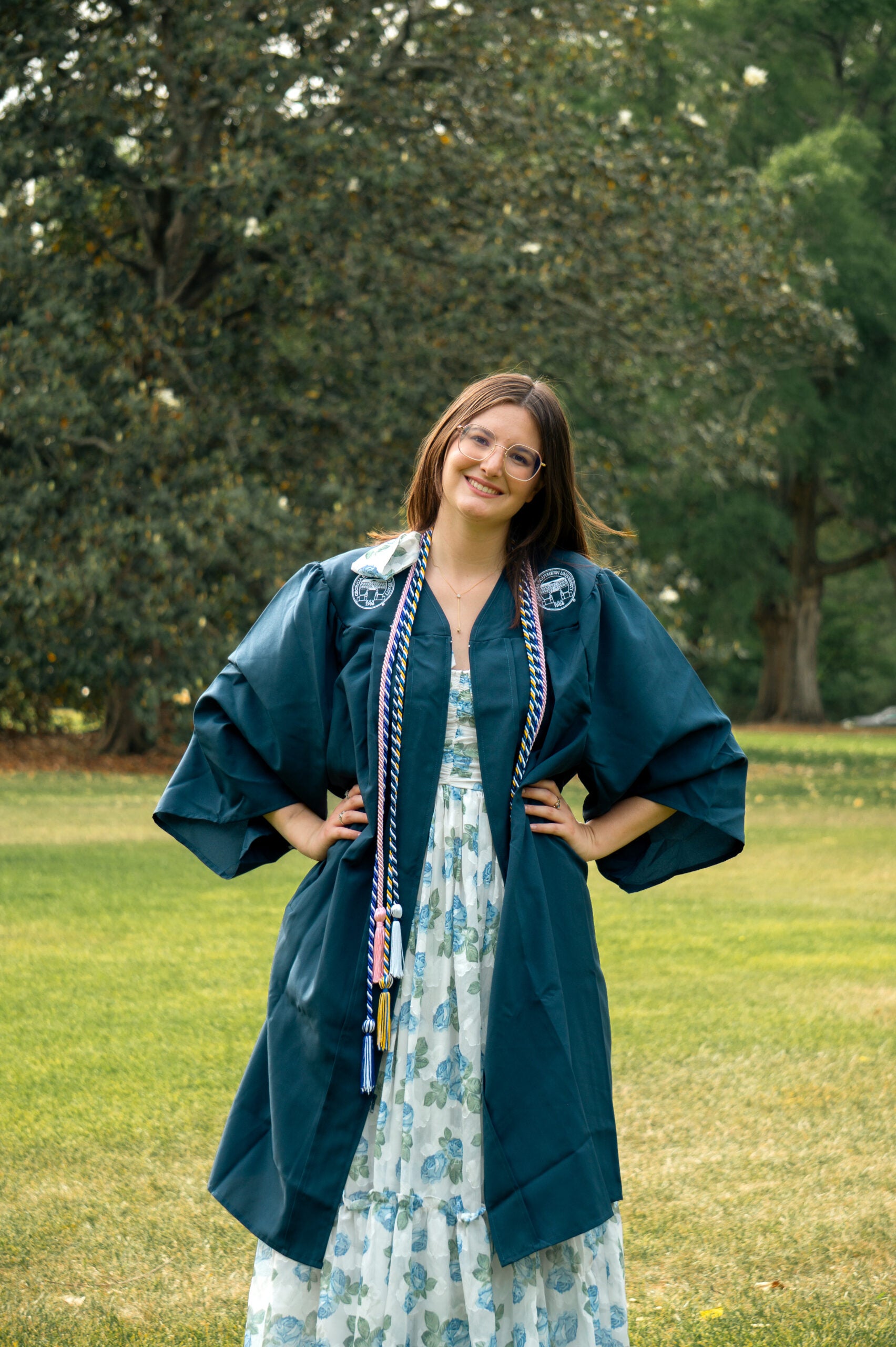 Graduate wearing teal cap and gown, smiling with hands on hips in an outdoor park.”,
