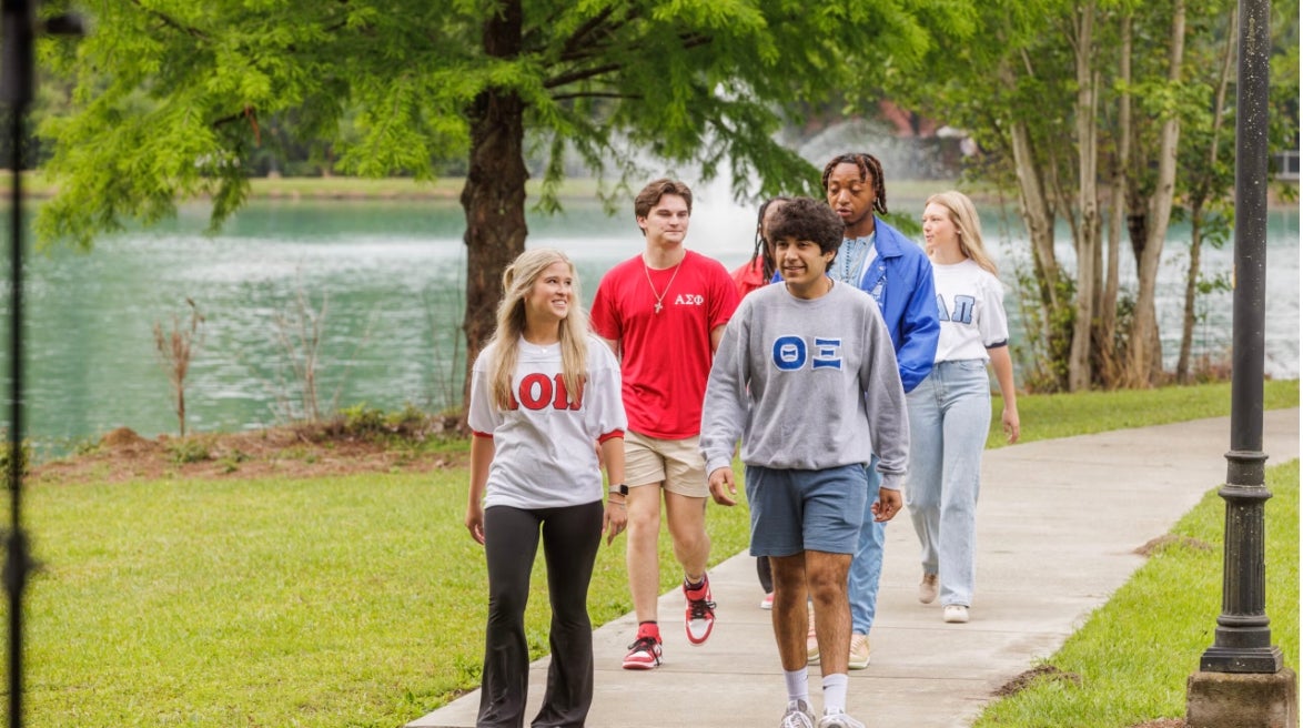 Group of five diverse young people walking together on a park path beside a lake, smiling.