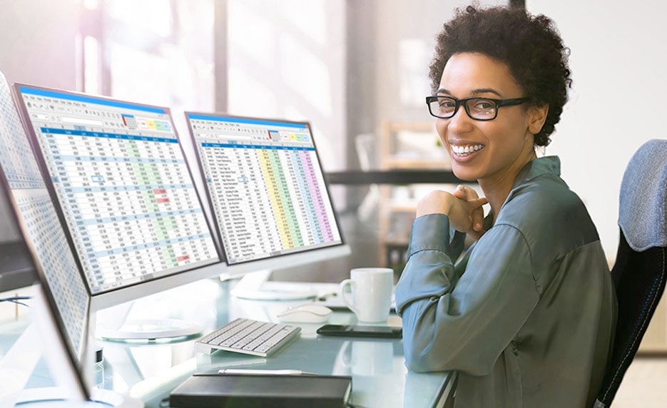 Woman with glasses sits at a desk with three monitors displaying spreadsheets, smiling at the camera.