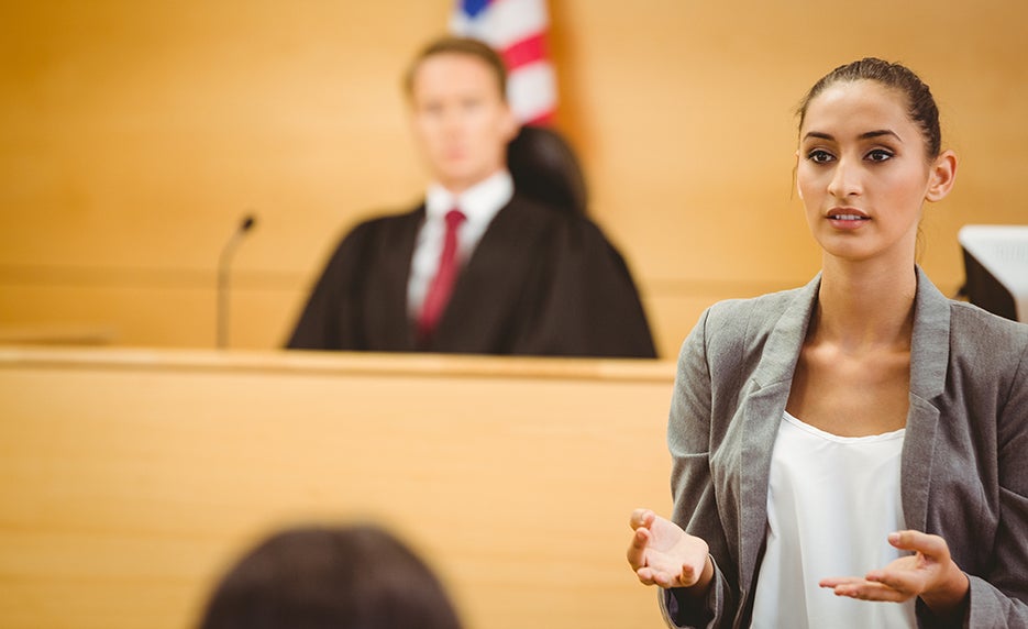Young female attorney in a gray blazer speaks to the courtroom, with a judge seated behind.