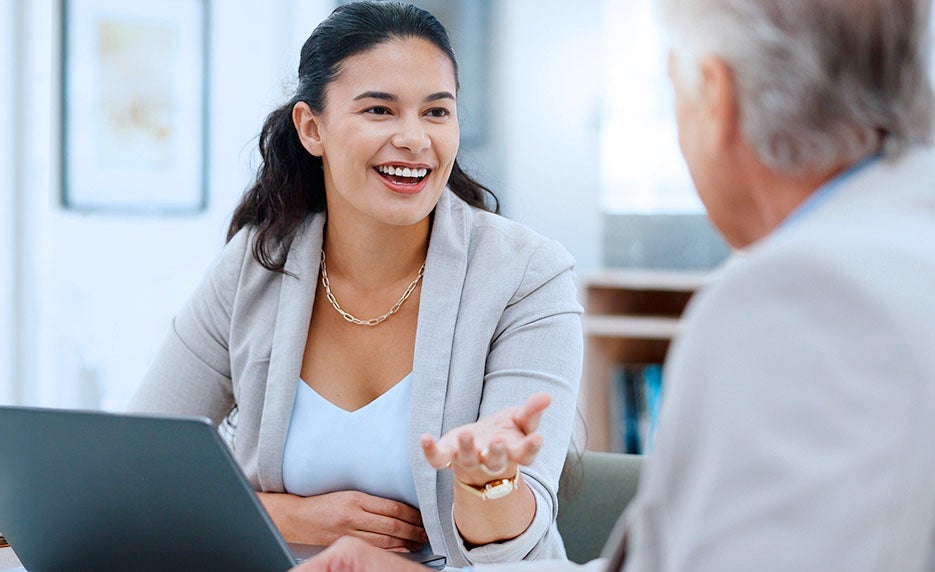 Professional woman smiling and gesturing while talking to an older man across a desk in a meeting room, laptop in view
