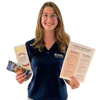 Smiling woman with glasses wearing a navy polo holds several nutrition pamphlets—one large folded sheet and two smaller booklets.
