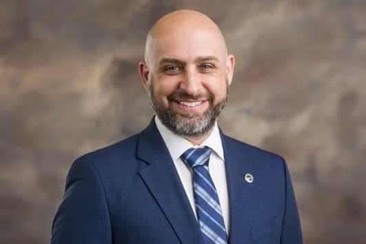Professional headshot of a smiling bald man in a blue suit and striped tie against a neutral brown backdrop, suitable for a corporate bio.
