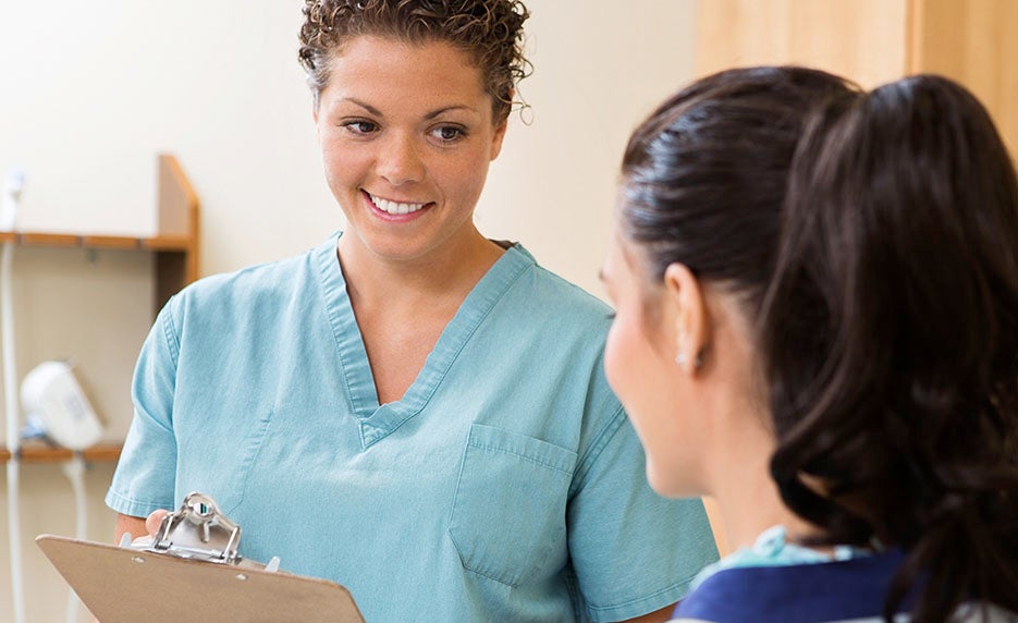 Nurse in blue scrubs holds a clipboard and smiles at a patient in a clinic examination room.