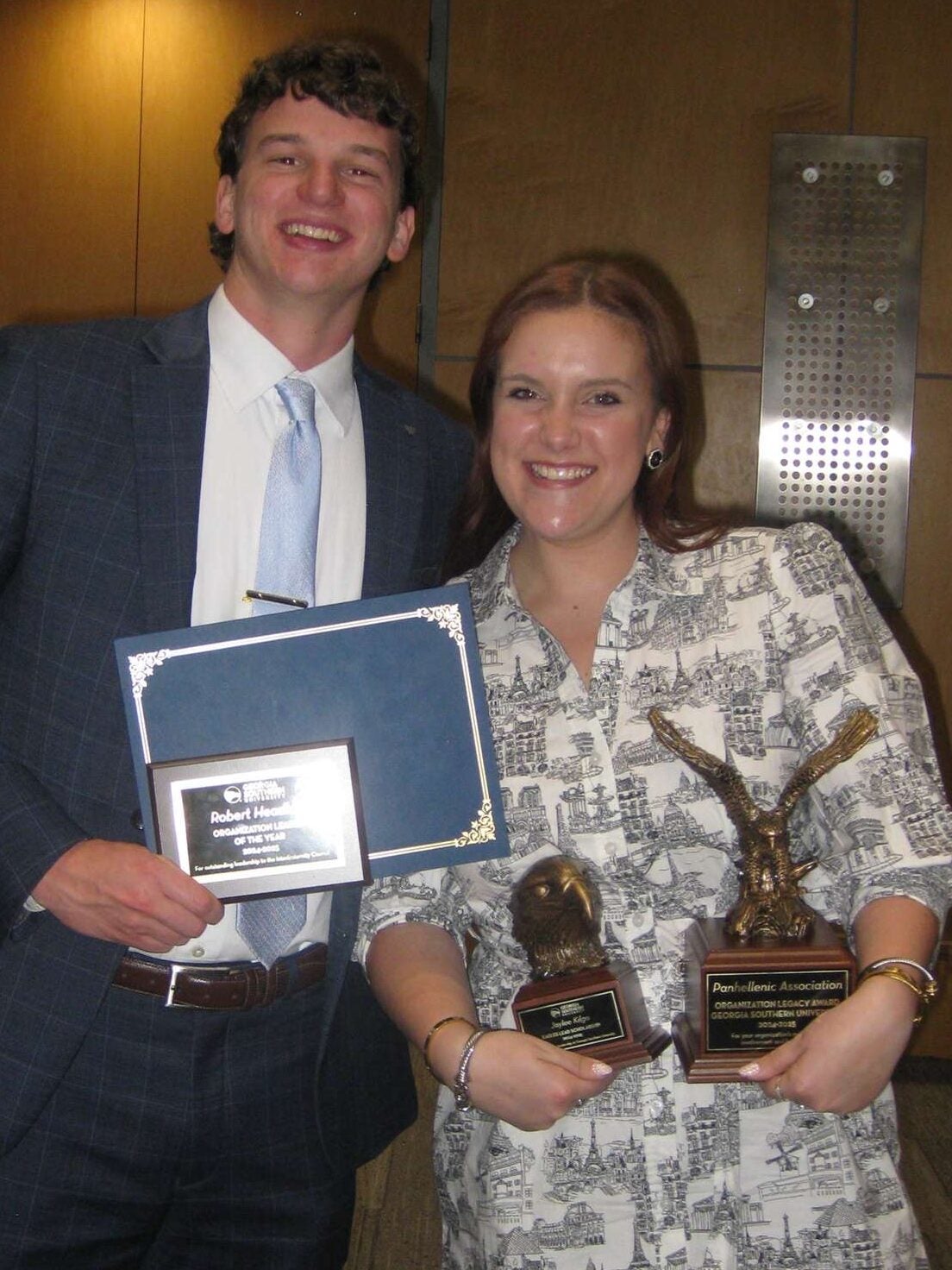 Two smiling people pose indoors with awards: a man in a blue suit holding a framed certificate and a plaque and a woman, Jaylee Kilgo, in a patterned dress holding two trophies.