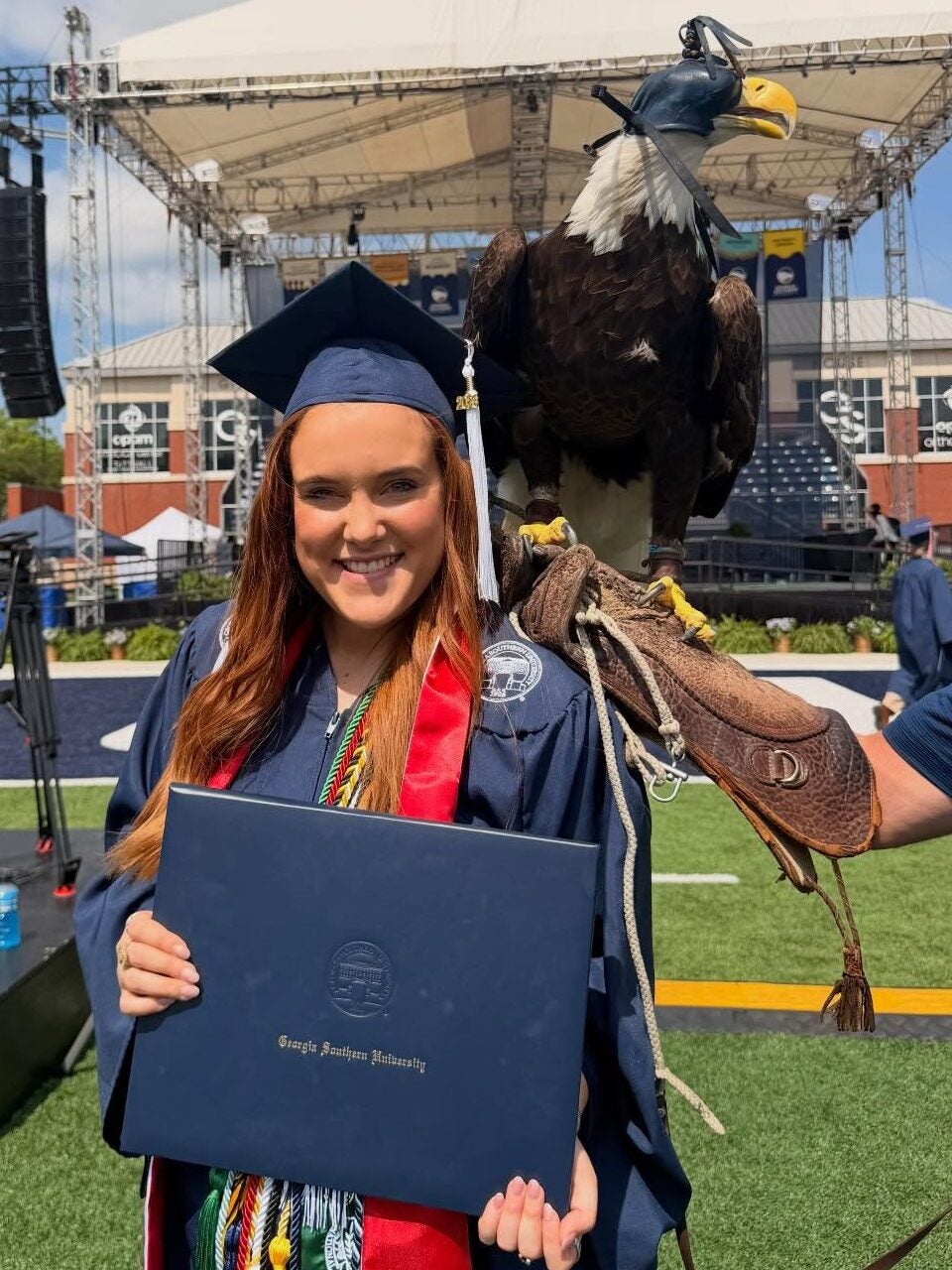 Jaylee Kilgo at commencement wearing Georgia Southern branded regalia and holding a diploma cover with Freedom II the eagle behind her shoulder.