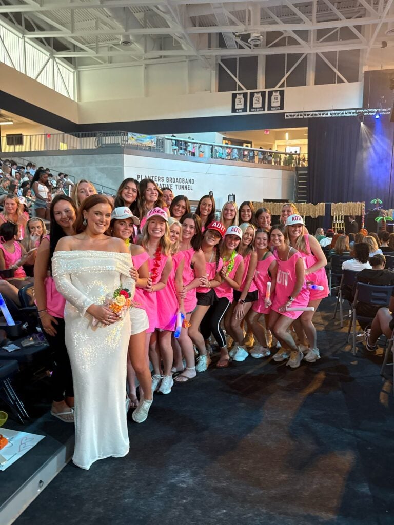 Jaylee Kilgo stands in a shimmering white dress surrounded by her sorority sisters who are wearing bright pink shirts.