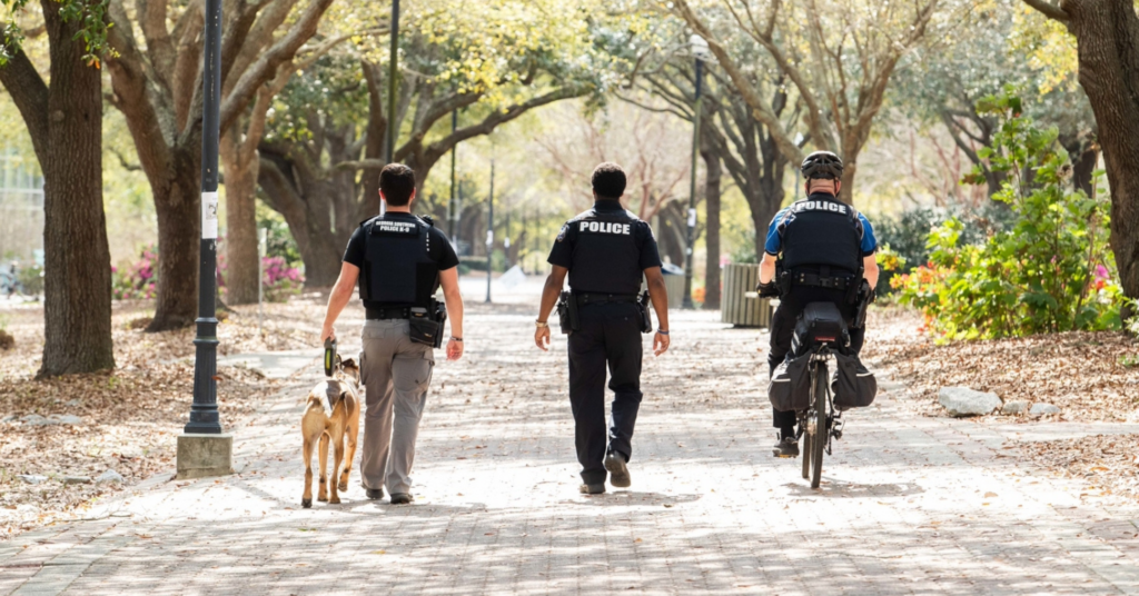 Three Georgia Southern Police Officers are moving away from the camera. The leftmost officer has a K-9, the center officer if walking and the rightmost officer is using a bicycle.