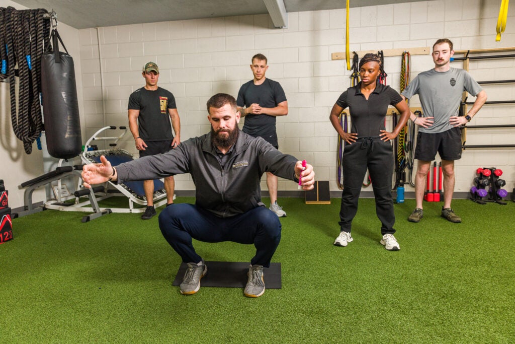 Bearded trainer performs a deep squat on a mat while four people stand watching in a gym.