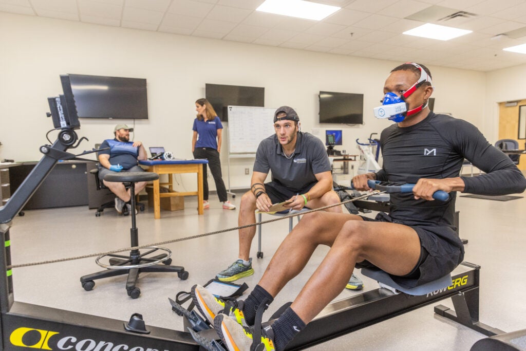 Athletes in a rehabilitation gym using stationary bikes while a trainer watches in the background.