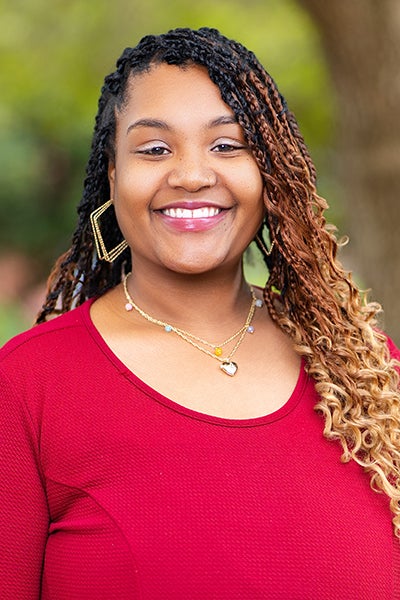Smiling woman posing outdoors in a red top with braided hair and gold earrings.