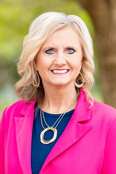 Smiling woman in a bright pink blazer outdoors, wearing gold hoop earrings and a circular pendant necklace.