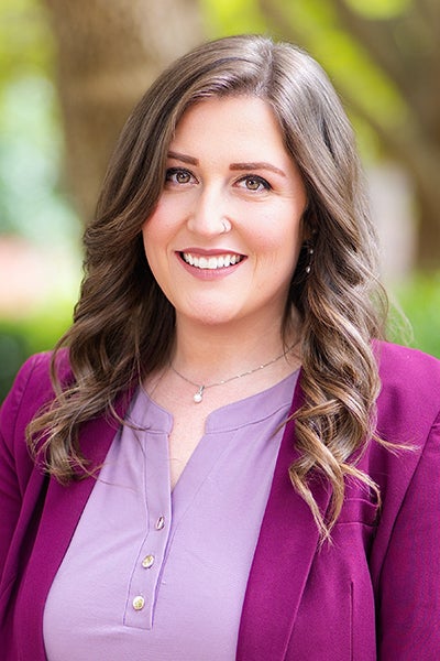 Portrait of a smiling woman with wavy brown hair, wearing a lavender blouse and magenta blazer, outdoors with greenery.
