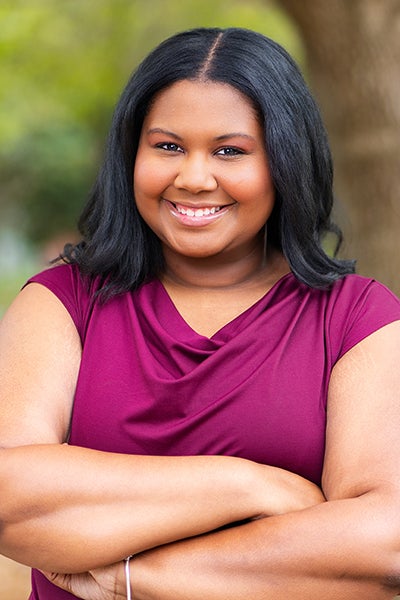 Smiling woman with arms crossed outdoors, wearing a magenta top.