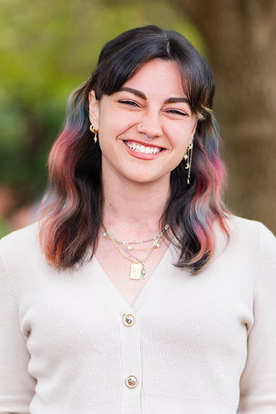 Smiling woman with dark hair tinted pastel pink and blue, wearing a beige cardigan and layered gold necklaces, outdoors.