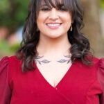 Smiling woman outdoors in a burgundy dress; dark hair with bangs and a leaf tattoo on her collarbone visible.