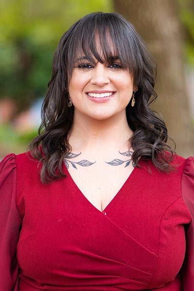 Smiling woman outdoors in a burgundy dress; dark hair with bangs and a leaf tattoo on her collarbone visible.