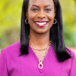 Professional head-and-shoulders portrait of a smiling woman in a magenta blazer with gold necklace, outdoors with a blurred green background