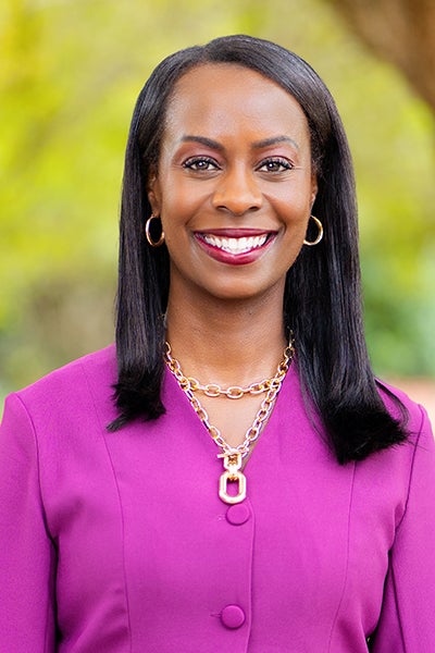 Professional head-and-shoulders portrait of a smiling woman in a magenta blazer with gold necklace, outdoors with a blurred green background