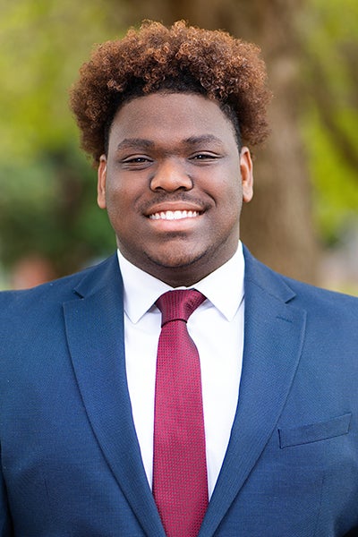 Smiling young man in a navy suit and red tie standing outdoors.