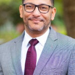 Man in a gray suit and burgundy tie, wearing glasses, smiling outdoors with greenery in the background.
