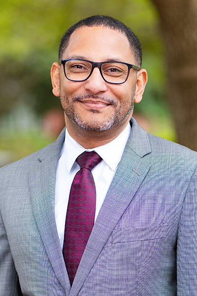 Man in a gray suit and burgundy tie, wearing glasses, smiling outdoors with greenery in the background.