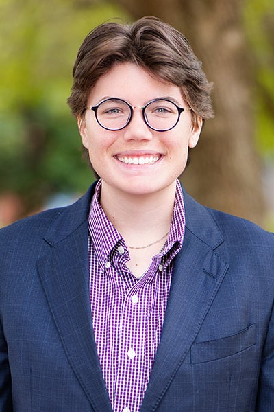 Smiling person in a blue blazer and purple checkered shirt outdoors with trees in the background.