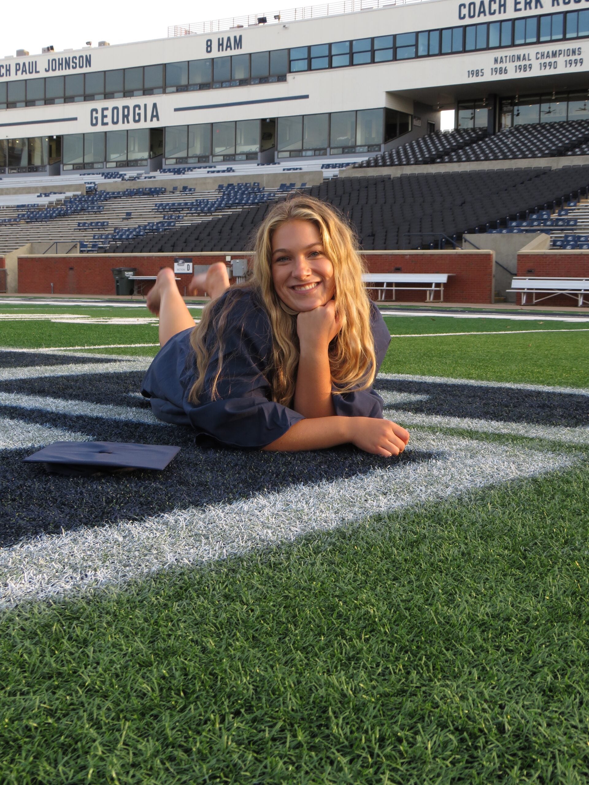 Smiling graduate in a navy gown lies on the football field with her cap beside her; stadium stands and a 'GEORGIA' sign are in the background.