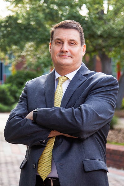 Businessman standing outdoors with arms crossed, wearing a navy suit and yellow tie, looking at the camera.