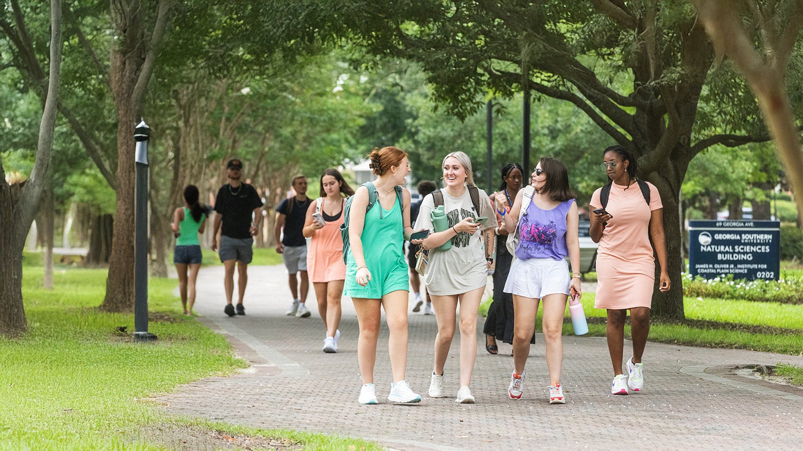 Group of diverse college students walking together along a tree-lined campus path and chatting.