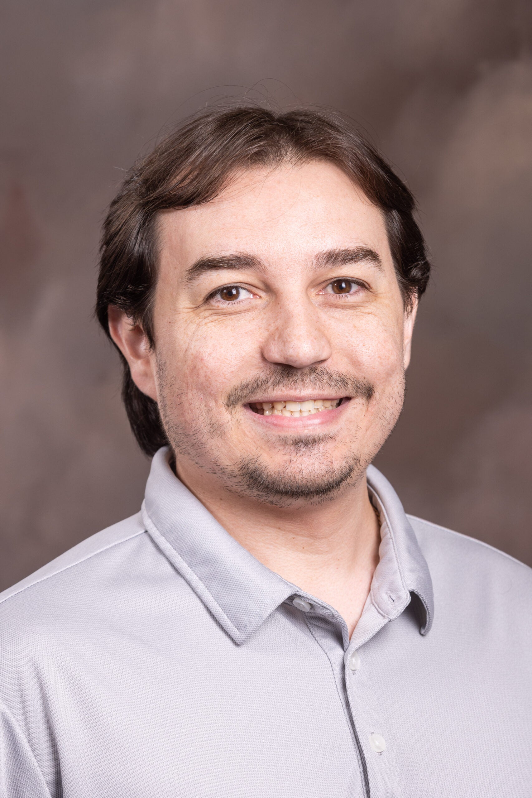 Close-up portrait of a smiling man with brown hair wearing a light gray button-down shirt against a brown backdrop.