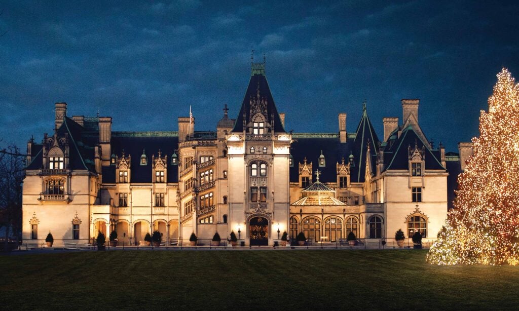 Biltmore Estates lit at dusk with a tall, brightly lit Christmas tree on the right.