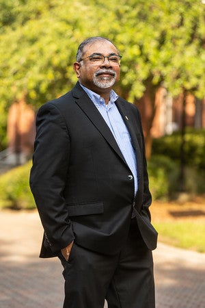 Dr. Abhijit "AB" Bhattacharyya, Dean is standing on campus. He is smiling and his hands in his pockets. He is wearing a navy blue suit and blue shirt. There is a walkway, trees, and a building in the background.