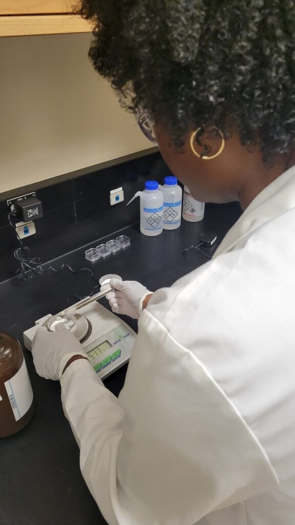 Scientist in a white lab coat and gloves weighs samples on a digital scale at a lab bench, with deionized water bottles in the background.