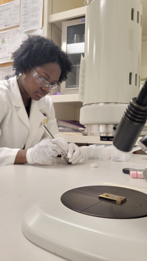 Scientist in a lab coat and safety goggles uses a tool at a workbench with a microscope nearby.