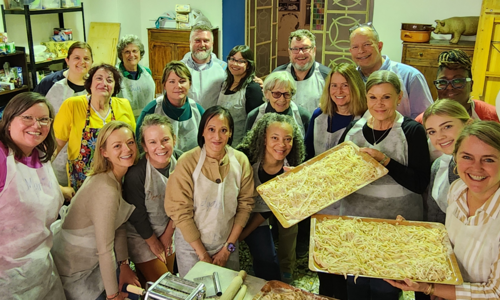 Group of CPE travelers in aprons posing with trays of prepared pasta in a bright kitchen.
