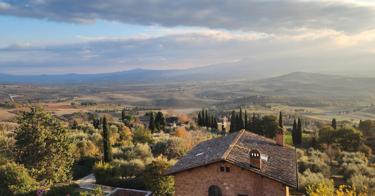 Sunlit Tuscan countryside with a stone house in the foreground, olive trees and cypress lining the landscape.