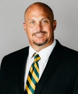 Professional head-and-shoulders portrait of a smiling man in a black suit and striped green‑yellow tie against a gray background.