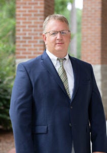 Man in a navy suit and striped tie posing outdoors beside brick columns.