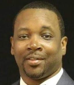 Close-up portrait of a man in a suit facing the camera, neutral expression on dark background.