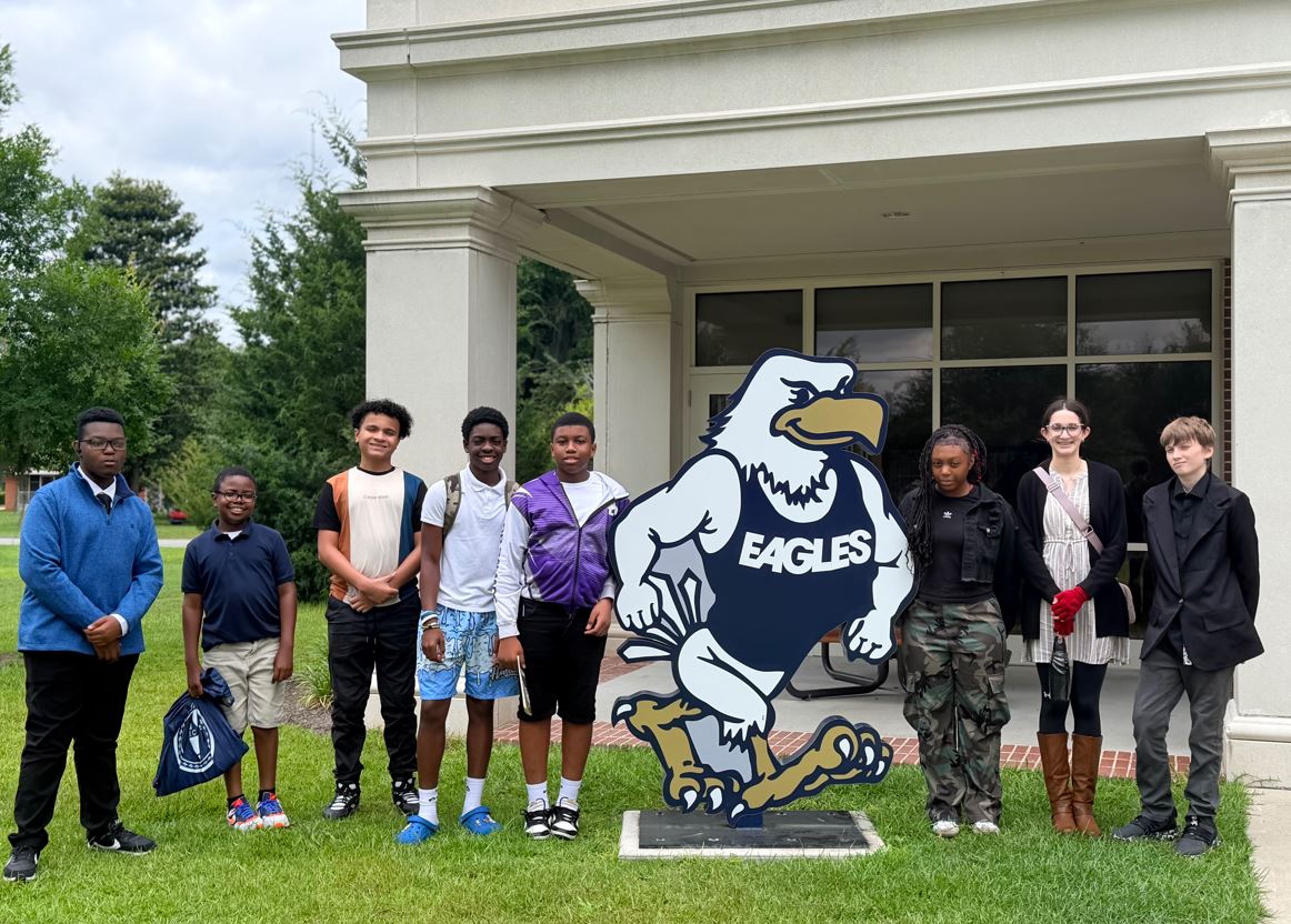 Group of diverse students standing on a grassy area beside a large cartoon eagle mascot outside a campus building.