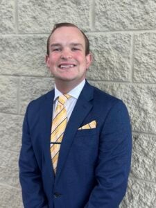 Portrait of a smiling man in a navy suit with a yellow striped tie, standing against a light gray brick wall with a pocket square showing.