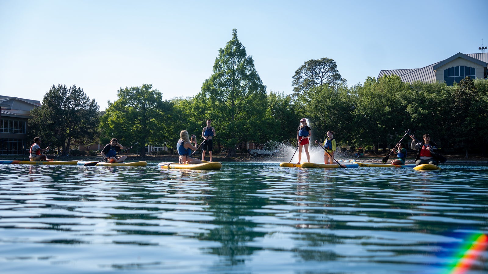 Group of six people stand-up paddleboarding on a calm lake with green trees and buildings along the shore in the background.
