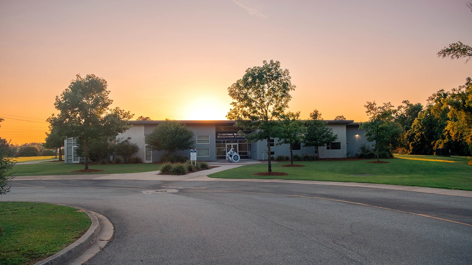 Shooting Sports Education Center building at sunset, with trees, manicured lawn, and a curved driveway in front.