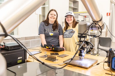 Two students are smiling and working in a manufacturing lab.