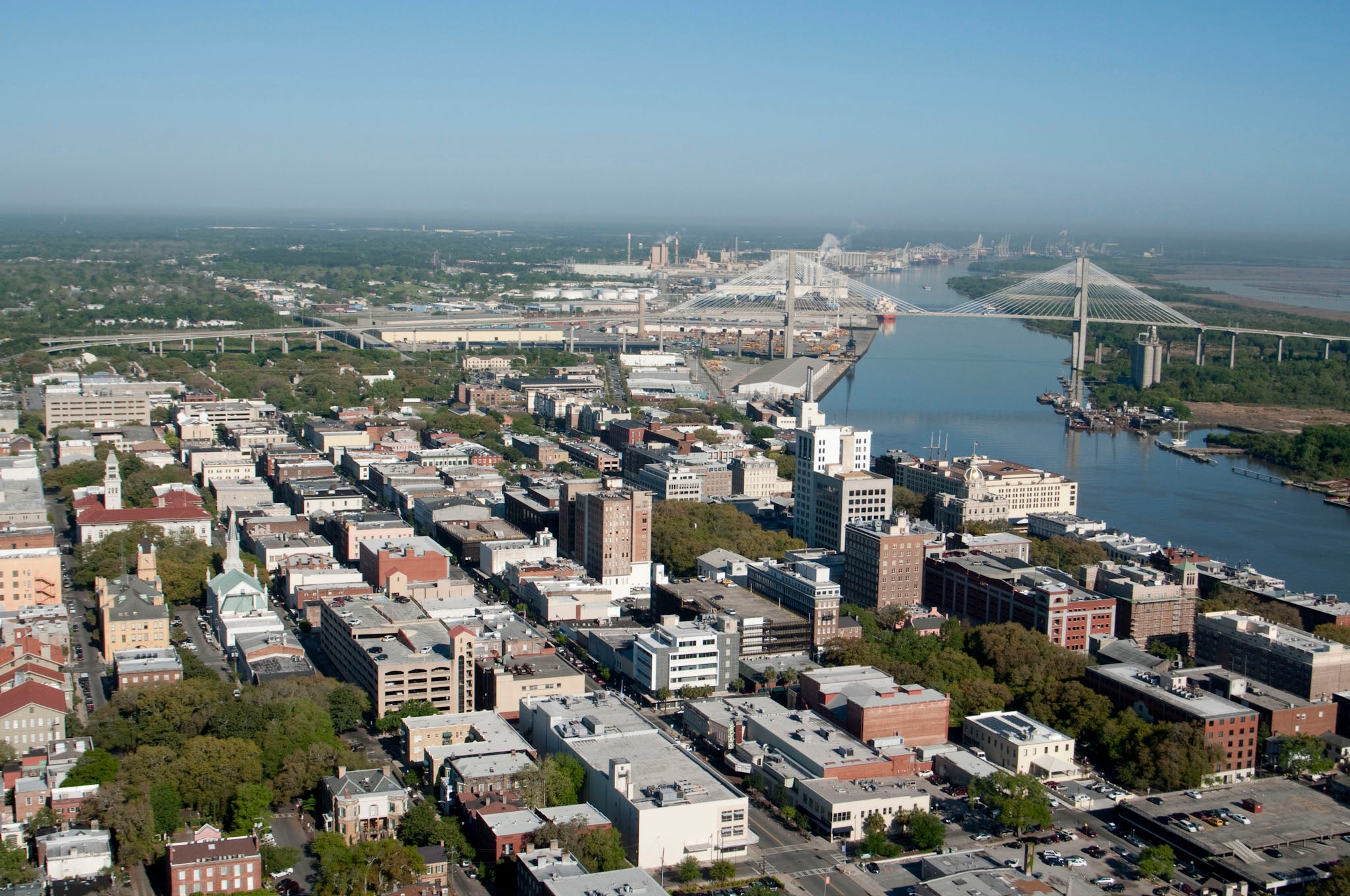 Aerial view of a city with a wide river and a cable-stayed bridge spanning it toward industrial port facilities in the distance.