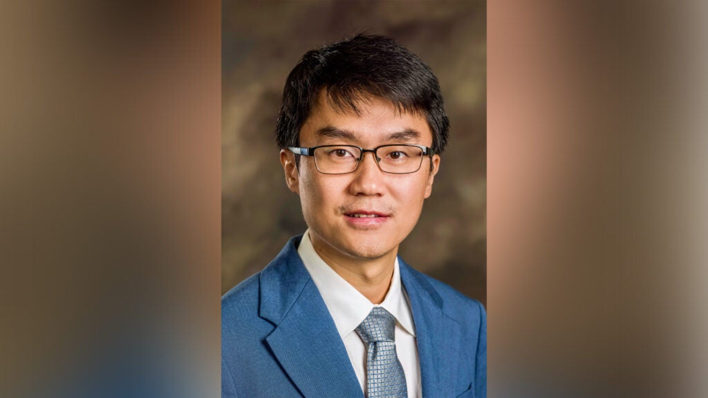 Portrait of an Asian man in a blue suit and tie, wearing glasses, looking at the camera with a neutral expression against a blurred brown backdrop.